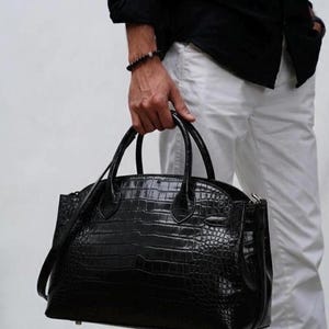 May include: A black leather handbag with a crocodile skin texture. The bag has two top handles and a shoulder strap. The bag is held by a person wearing a black shirt and white pants.