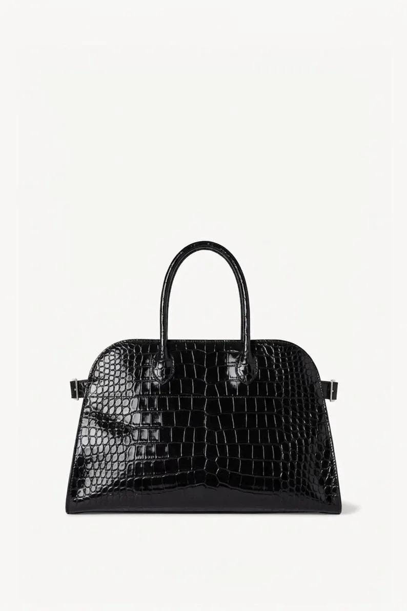 May include: Black, glossy handbag with a textured, crocodile-like pattern. The bag has a curved top, a short handle, and small metal accents on the sides. The bag is set against a white background.