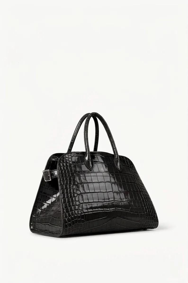May include: A black, structured handbag with a crocodile-skin texture. The bag has two curved handles and a silver-tone buckle closure. The bag is a trapezoid shape and sits on a white background.