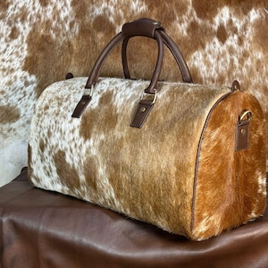 May include: A cowhide leather duffel bag with brown leather handles and accents. The bag has a natural brown and white spotted pattern. The bag is cylindrical with a zip closure and a handle on each end. The bag is on a brown leather surface.