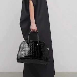 May include: A black, glossy handbag with a crocodile-skin texture. The bag has a rounded shape with a top handle and is held by a person wearing a black dress.