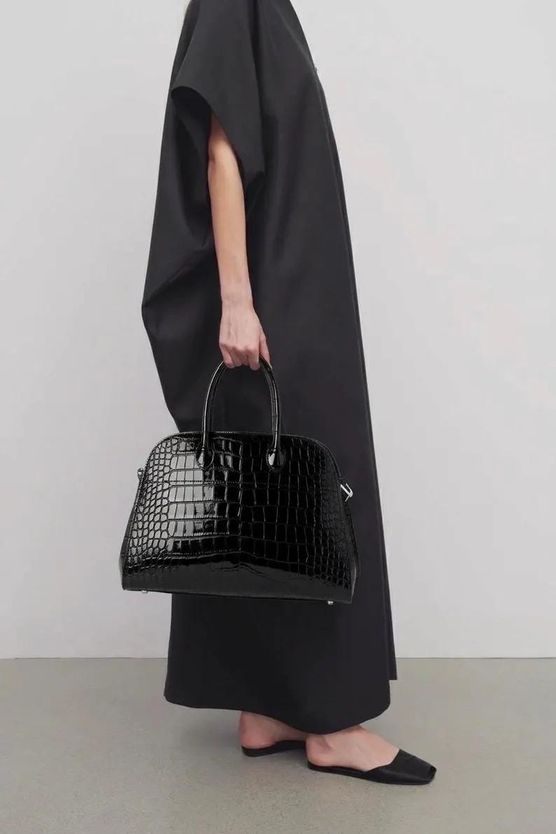 May include: A black, glossy handbag with a crocodile-skin texture. The bag has a rounded shape with a top handle and is held by a person wearing a black dress.