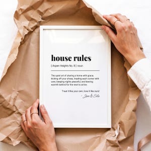May include: A white framed wall art print featuring the text house rules, Aspen Heights No. 8, and a definition about sharing a home with grace. The print includes the phrase Treat it like your own, love it like ours! and is signed by Jon & Sila.