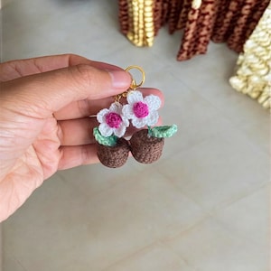 May include: Crocheted flower earrings with pink centers and green leaves, hanging from gold hoops. The flowers are in brown pots.