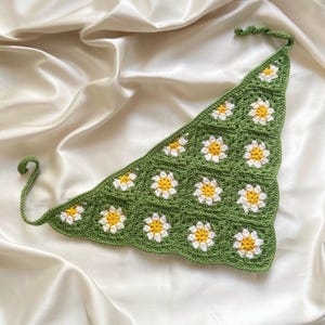 May include: A triangular crochet bandana featuring a repeating pattern of daisy motifs. The design consists of green square panels, each containing a white and yellow flower. The accessory includes two thin crocheted ties at the top and side corners.