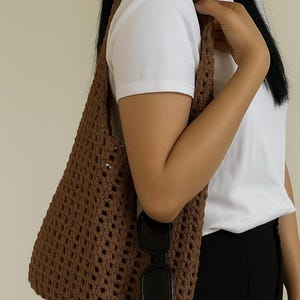 May include: A brown, open-weave crochet tote bag with a shoulder strap. The bag is held by a person wearing a white t-shirt and black pants. Black sunglasses are tucked into the bag.