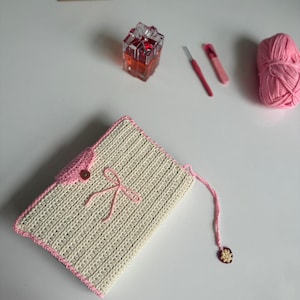 May include: A handmade, cream-coloured crochet clutch with a pink trim, button, and bow. A pink yarn pull with a decorative charm extends from the clutch. Other items include pink yarn, crochet hooks, and a small framed floral artwork.