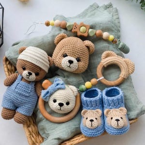 May include: A collection of handmade crochet baby items, including a teddy bear doll in blue overalls, a bear head, a bear-shaped teether, a pacifier clip, and blue booties with bear faces. The items are in a wicker basket on a green cloth.