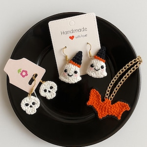 May include: A collection of handmade Halloween-themed crochet jewelry. Includes white ghost earrings with black hats, skull earrings, and an orange bat necklace. The earrings and necklace are displayed on a black plate. The text "Handmade with love" is visible.