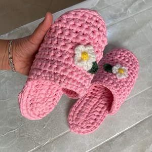 May include: A pair of pink crocheted slippers with white flower embellishments. The slippers are made with a soft, textured yarn and have a comfortable, open-toe design.