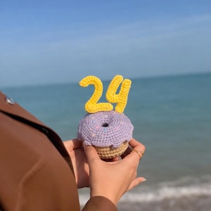 Op de afbeelding: Een gehaakte cupcake met een lichtpaarse bovenkant, een bruine basis en het gele nummer 24 erop. De cupcake wordt vastgehouden tegen een blauwe oceaanachtergrond.
