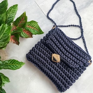 May include: A dark blue crocheted crossbody bag with a gold clasp. The bag has a chain strap.