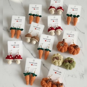 May include: A selection of handmade crochet earrings in various designs. The earrings include orange carrots with green tops, red and white mushroom caps, brown mushrooms, and orange pumpkins. Each pair is displayed on a white card with the text "Handmade with love."
