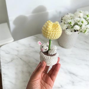 May include: A crocheted white bunny sitting in a white pot with a yellow crocheted flower growing out of the pot.