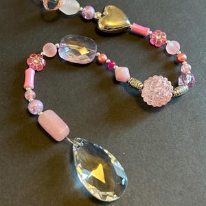 May include: A pink and silver beaded suncatcher with a crystal teardrop pendant. The beads are various shapes and sizes, including flowers, hearts, and round beads. The suncatcher is hanging from a silver ring.