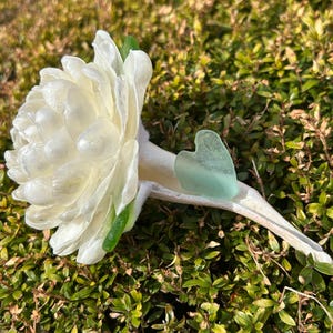 May include: A white flower with a green stem and a light blue heart-shaped piece of sea glass resting inside the flower.