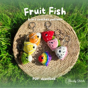 May include: A collection of six colorful crochet fruit fish keychains, each with a silver keyring. The keychains are arranged on a woven, circular mat, with the text "Fruit Fish" and "6-in-1 crochet pattern" in white. The words "PDF download" are also visible.