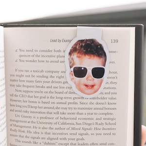 Può includere: Primo piano di un libro con un segnalibro che mostra un bambino con occhiali da sole. Il titolo del libro è "Lead by Example" e il numero di pagina è 139. Il testo della pagina tratta dell'importanza di allineare gli incentivi con gli obiettivi organizzativi.