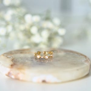 Könnte beinhalten: Ein goldfarbener offener Ring mit filigranem Design. Der Ring hat kleine, runde, weiße Edelsteine und einen winzigen, klaren Edelstein in der Mitte. Der Ring wird auf einer hellen, oval geformten Steinoberfläche präsentiert.