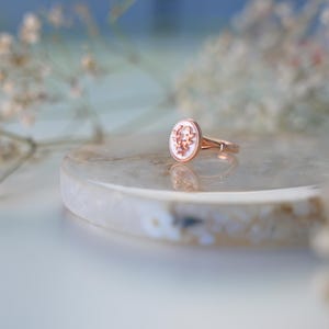 May include: A rose gold ring with an oval, white cameo featuring a floral design. The ring is set on a light-coloured, round stone surface, with soft focus flowers in the background.