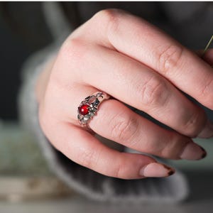 May include: A silver ring with an ornate design, featuring a round, red gemstone at its centre. The ring is worn on a finger, showcasing its intricate details and the vibrant colour of the stone. The ring's design is floral and delicate.
