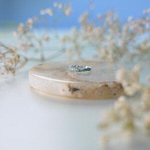 Op de afbeelding: Een zilverkleurige ring met turquoise accenten ligt op een lichtbruine, ovale steen. De ring is het middelpunt, met delicate witte bloemen en een zachte blauwe achtergrond. De ring is een sieraad.