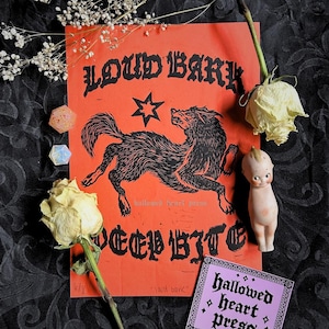 May include: An orange print with a black wolf illustration and the text "LOUD BARK DEEP BITE". A small doll, dried roses, and a purple sticker with the text "hallowed heart press" are also present.