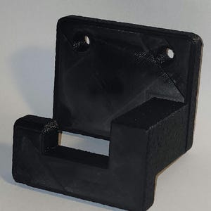 May include: A black plastic wall mount with a single hook. The hook is designed to hold a small object, such as a phone or a set of keys.