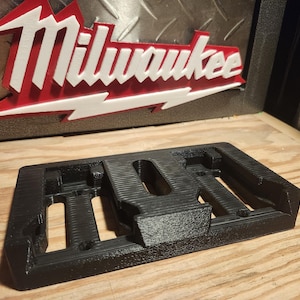 Puede incluir: Organizador de plástico negro impreso en 3D con cuatro ranuras y un compartimento central. El organizador está diseñado para guardar objetos pequeños como herramientas o baterías.