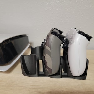 May include: A black charging stand with two video game controllers. One controller is white and the other is camouflage.