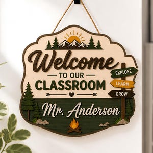May include: A decorative wooden wall sign featuring the text Welcome to our classroom and Mr. Anderson. The design includes a sun, mountains, pine trees, a campfire, and three directional signs labeled Explore, Learn, and Grow, all suspended by a rope.