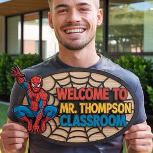 May include: An oval wooden sign with a Spider-Man graphic and the words "WELCOME TO MR. THOMPSON CLASSROOM" in red, yellow, and blue. The sign has a spiderweb design and is held up by a person.