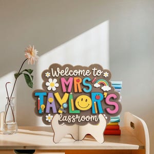 May include: A wooden sign with the text "Welcome to MRS TAYLOR'S classroom" in colorful letters. The sign features a smiley face, a rainbow, and other school-themed icons. It is displayed on a wooden stand.
