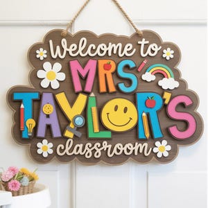 May include: A wooden classroom sign with the text "Welcome to MRS TAYLOR'S classroom." The sign features colorful letters, a smiley face, daisies, and school-themed illustrations like pencils and an apple. It hangs from a jute rope.