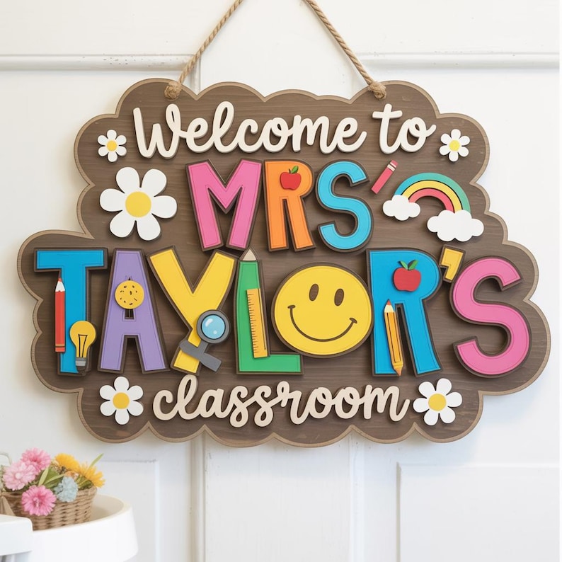May include: A wooden classroom sign with the text "Welcome to MRS TAYLOR'S classroom." The sign features colorful letters, a smiley face, daisies, and school-themed illustrations like pencils and an apple. It hangs from a jute rope.