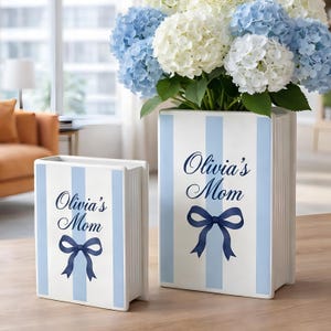 Puede incluir: Dos jarrones de cerámica blanca con rayas azules y el texto "Olivia's Mom" y un lazo azul. El jarrón más grande contiene hortensias azules y blancas. Los jarrones están sobre una mesa de madera.