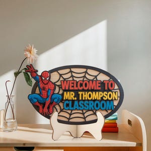 May include: Wooden classroom sign featuring Spider-Man and the text "WELCOME TO MR. THOMPSON CLASSROOM". The sign is oval-shaped with a black web design and a wooden stand. The sign is displayed on a wooden surface.