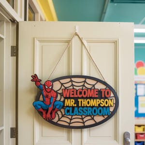 May include: Oval wooden classroom sign with a Spider-Man graphic and the text "WELCOME TO MR. THOMPSON CLASSROOM." The sign is hanging on a white door with a beige rope.