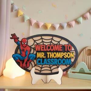 May include: A wooden classroom sign featuring Spider-Man. The oval sign has a black web design and the words "WELCOME TO MR. THOMPSON CLASSROOM" in red, yellow, and blue. The sign is supported by a white wooden stand.