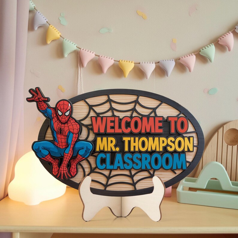 May include: A wooden classroom sign featuring Spider-Man. The oval sign has a black web design and the words "WELCOME TO MR. THOMPSON CLASSROOM" in red, yellow, and blue. The sign is supported by a white wooden stand.