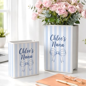 Puede incluir: Dos jarrones de cerámica blanca con rayas verticales azules. Los jarrones están inscritos con "Chloe's Nana" y una ilustración de lazo. Un jarrón contiene un ramo de rosas rosas. Los jarrones están sobre una superficie blanca.