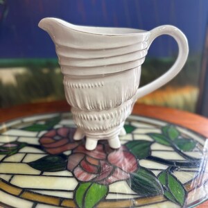 May include: A white ceramic creamer with a unique, textured design. The creamer has a spout, a handle, and rests on three small feet. The body features horizontal ridges and a decorative pattern. The creamer is displayed on a decorative glass surface.