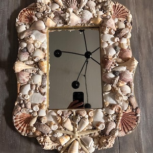 May include: A rectangular mirror with a gold frame, intricately decorated with a variety of seashells. The frame is adorned with numerous shells in shades of white, beige, and brown, creating a textured, coastal-themed design.