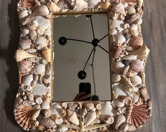 May include: A rectangular mirror with a gold frame, intricately decorated with a variety of seashells. The frame is adorned with numerous shells in shades of white, beige, and brown, creating a textured, coastal-themed design.