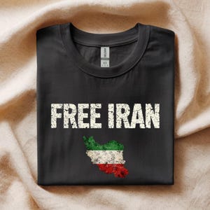 以下が含まれることがあります： 黒いTシャツ。白の擦り切れた文字で「FREE IRAN」と書かれています。テキストの下には、イランの国旗の色（緑、白、赤）でイランの地図のグラフィックがあります。シャツはベージュ色の布の上に折りたたまれています。