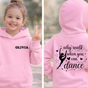 May include: Pink hooded sweatshirt with the name "OLIVIA" printed in black on the front. The back features a ballerina graphic and the text "why walk when you can dance" in black. The sweatshirt has a front pocket.