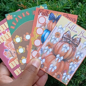 Può includere: Una mano tiene quattro carte colorate a tema autunnale. Le carte presentano foglie autunnali, zucche e fiocchi. Il testo sulle carte include "Leaves", "Fall" e "Fallin' Bows". Le carte hanno un design decorativo con colori autunnali.