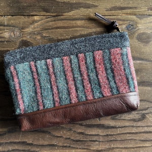 May include: A striped wool zippered pouch with a brown leather bottom. The pouch has a gray, pink, and green striped pattern.