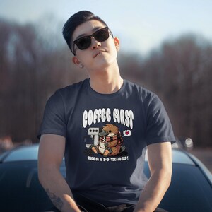 May include: A navy blue t-shirt with a cartoon platypus holding a cup of coffee. The text "Coffee First, Then I Do Things!" is printed on the shirt.