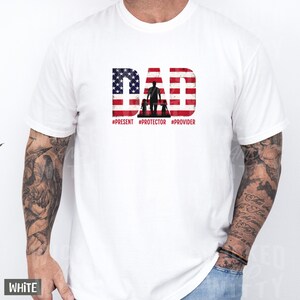 May include: White t-shirt with a graphic design featuring the word "DAD" in red and blue, resembling the American flag. The design includes a silhouette of a person with two children and the hashtags #PRESENT #PROTECTOR #PROVIDER.